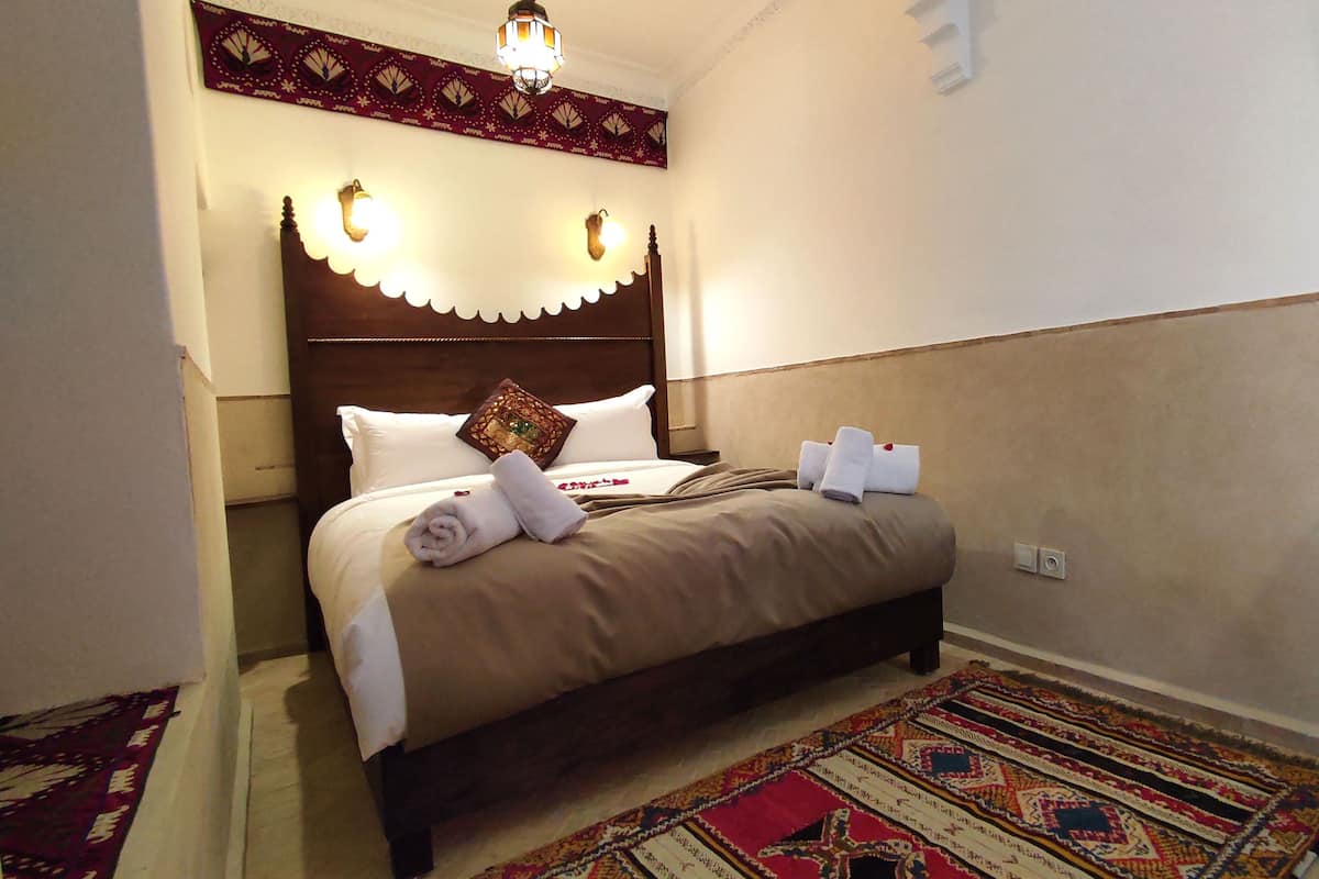 A neatly made bed with towels and a decorative pillow, a patterned rug, and a wall-mounted light fixture.