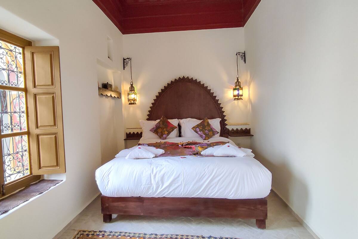 A neatly made bed with a wooden headboard, two wall-mounted lamps, and a window with decorative iron bars.