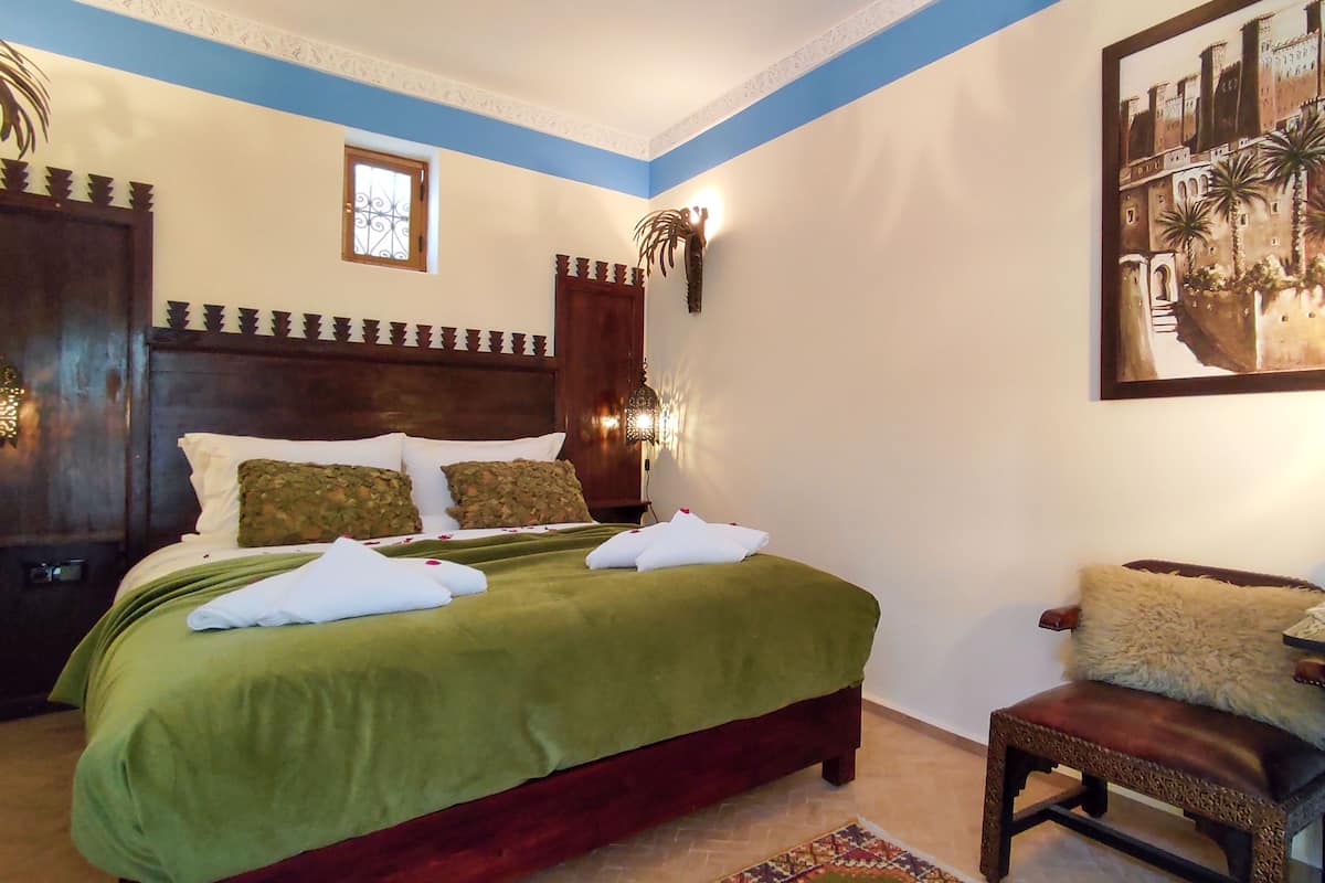 A neatly made bed with a green bedspread and white pillows, a wooden headboard, a wall-mounted lamp, and a framed painting of a building.