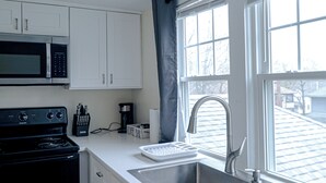 A kitchen with white cabinets, a microwave, a stove, a coffee maker, and a window with a view of a house. - 79-4 spacious studio Walk to backus hospital and downtown (Norwich)