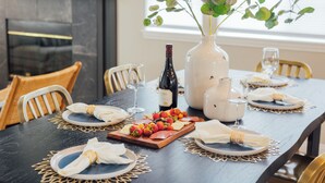 Una mesa de comedor dispuesta con platos, servilletas, una botella de vino, un florero con follaje y una fuente de frutas.