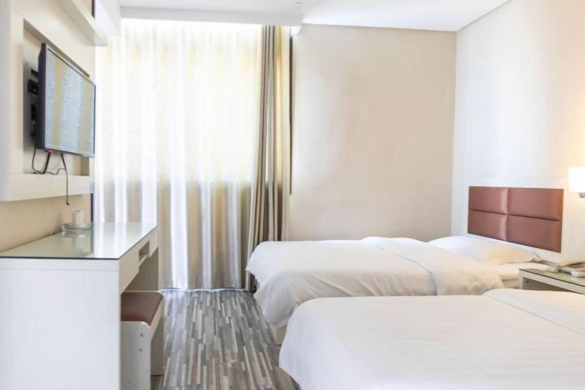 A hotel room with two beds, a desk, a TV mounted on the wall, and a window with curtains.