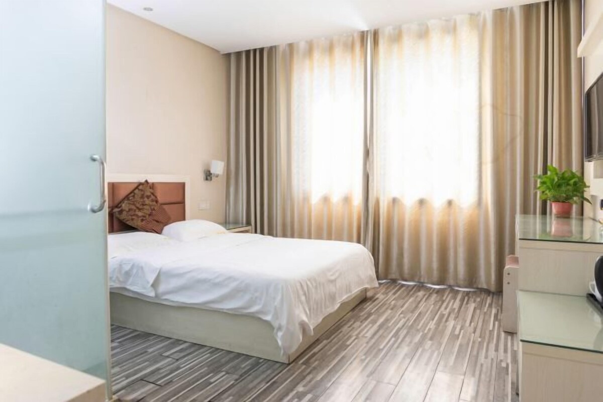 A hotel room with a bed, a desk, a chair, a potted plant, and a window with curtains.