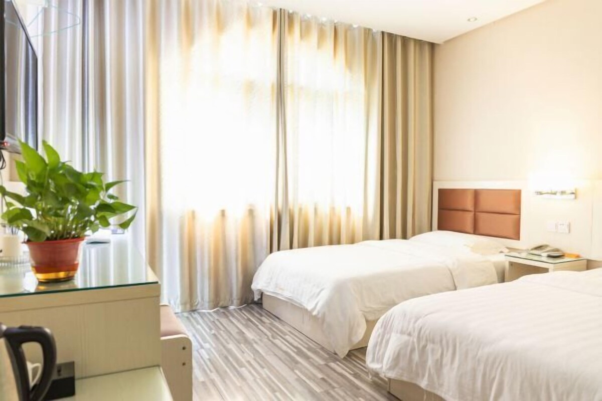 A hotel room with two beds, a desk, a chair, a potted plant, and a window with curtains.