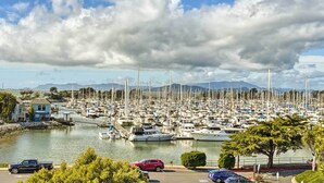 A marina with numerous boats docked, a parking area with cars, and a backdrop of mountains and a partly cloudy sky.