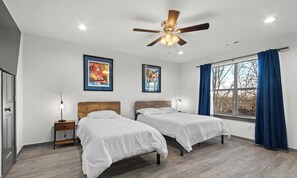 A hotel room with two beds, a ceiling fan, artwork on the walls, a window with blue curtains, and a wooden floor.