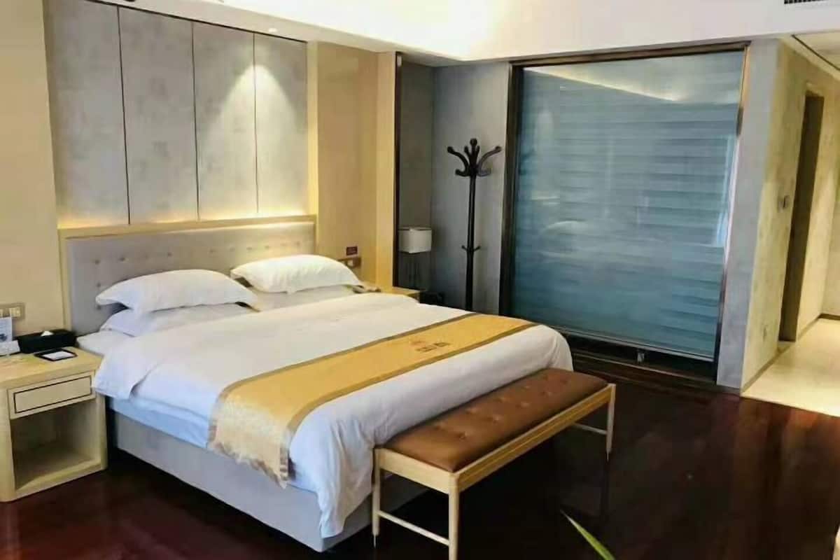 A hotel room with a bed, bedside table, bench, coat rack, and a large window.
