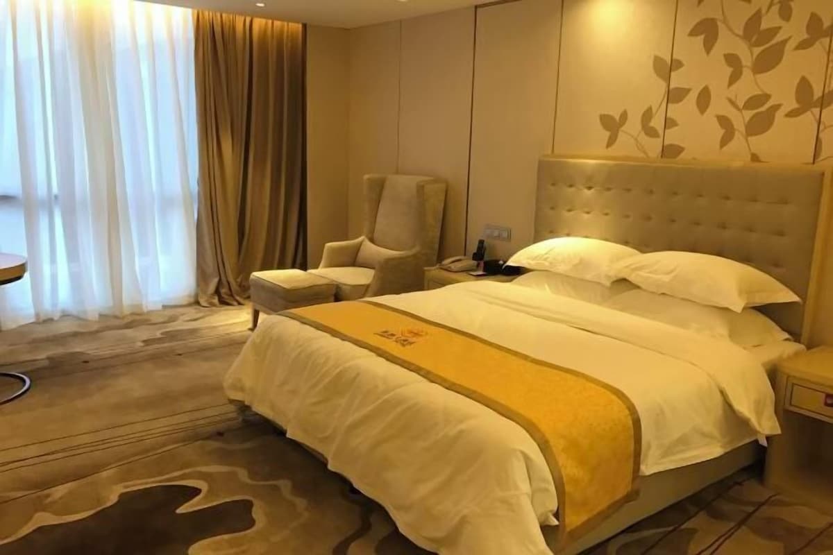 A hotel room with a bed, a chair, a bedside table, and a large window with curtains.