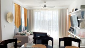 Un salon moderne avec un coin repas, comprenant un ventilateur de plafond, un ensemble de canapés, un téléviseur posé sur une étagère blanche et un tapis à motifs.