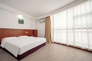 A hotel room with a large bed, a wooden headboard, a nightstand, a window with sheer curtains, and a painting on the wall.