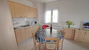 A compact kitchen with wooden cabinets, a dining table with chairs, a TV, and a window with curtains. - Apartment Branka for 2 -3 person (Premantura)