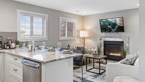 A modern kitchen with a built-in dishwasher, a dining area with a fireplace, and a flat-screen TV mounted above the fireplace.