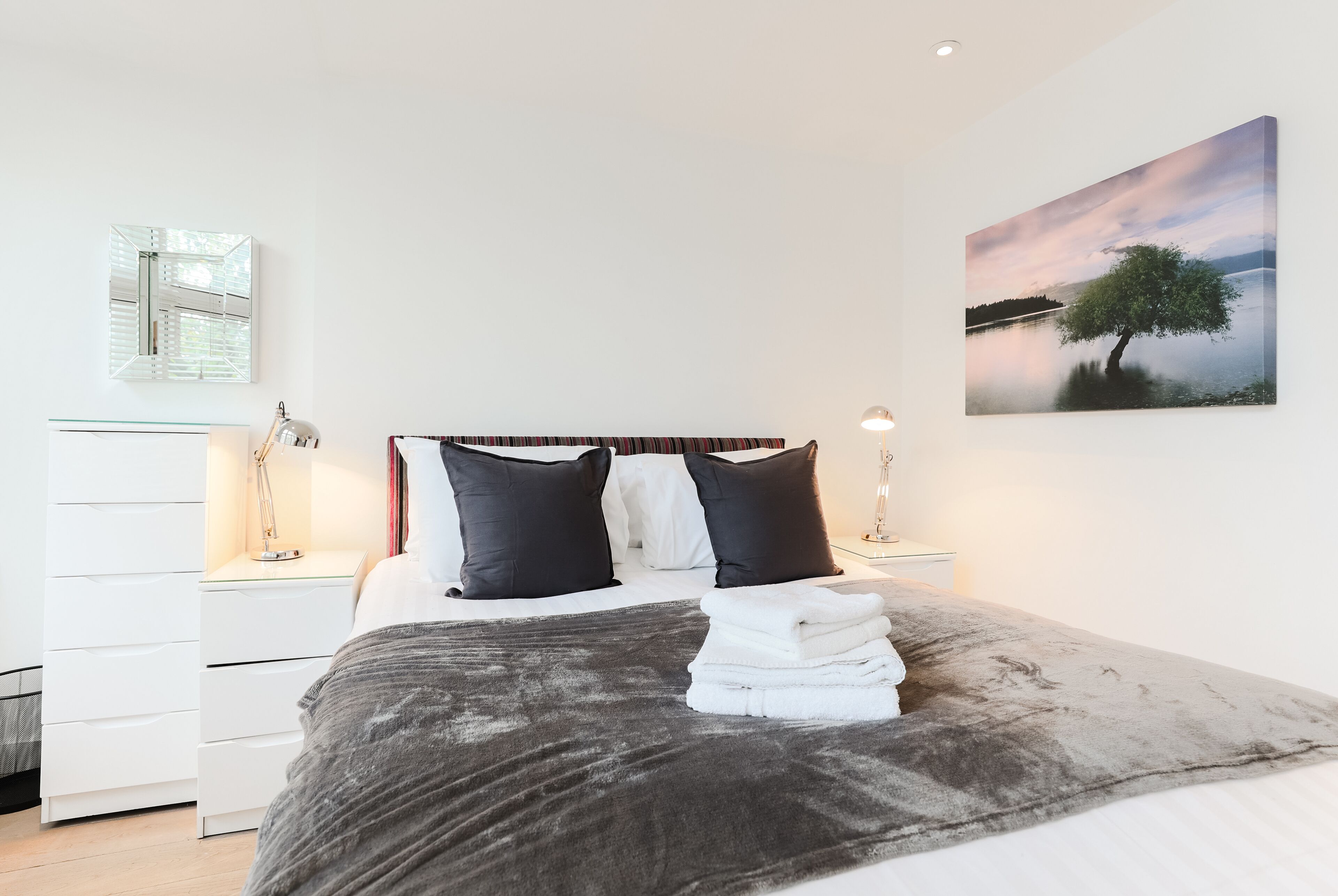 Una cama bien hecha con una manta gris, dos almohadas negras y una pila de toallas blancas. En la pared, un cuadro de un árbol junto a un lago.