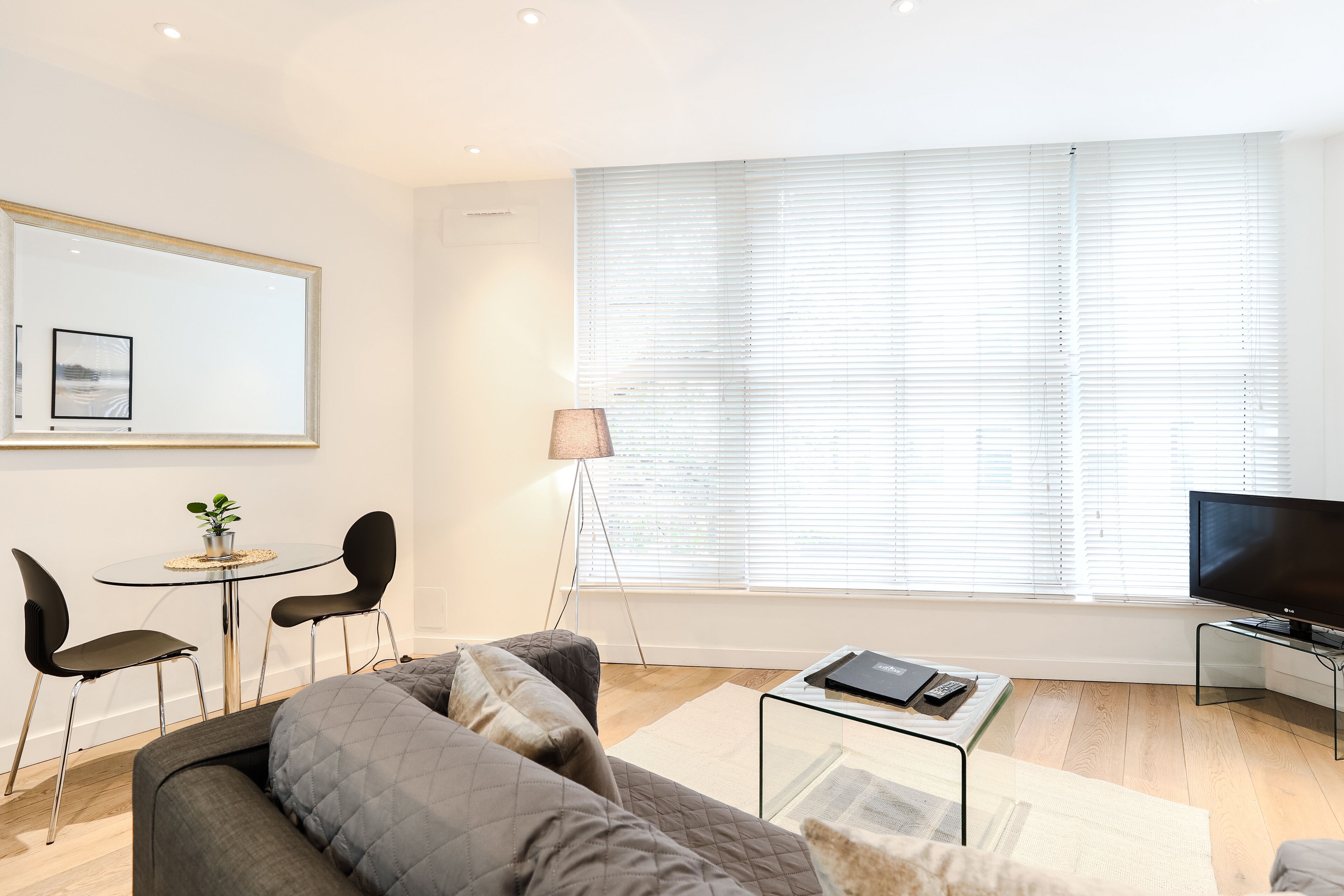 A modern living room with a grey sofa, a small round table with two chairs, a floor lamp, a flat-screen TV, and a large window with blinds.