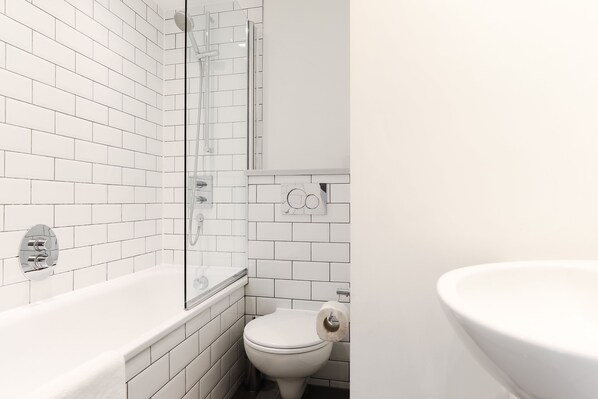 A bathroom with white tiled walls, a glass shower enclosure, a white bathtub, and a toilet with a paper towel holder.