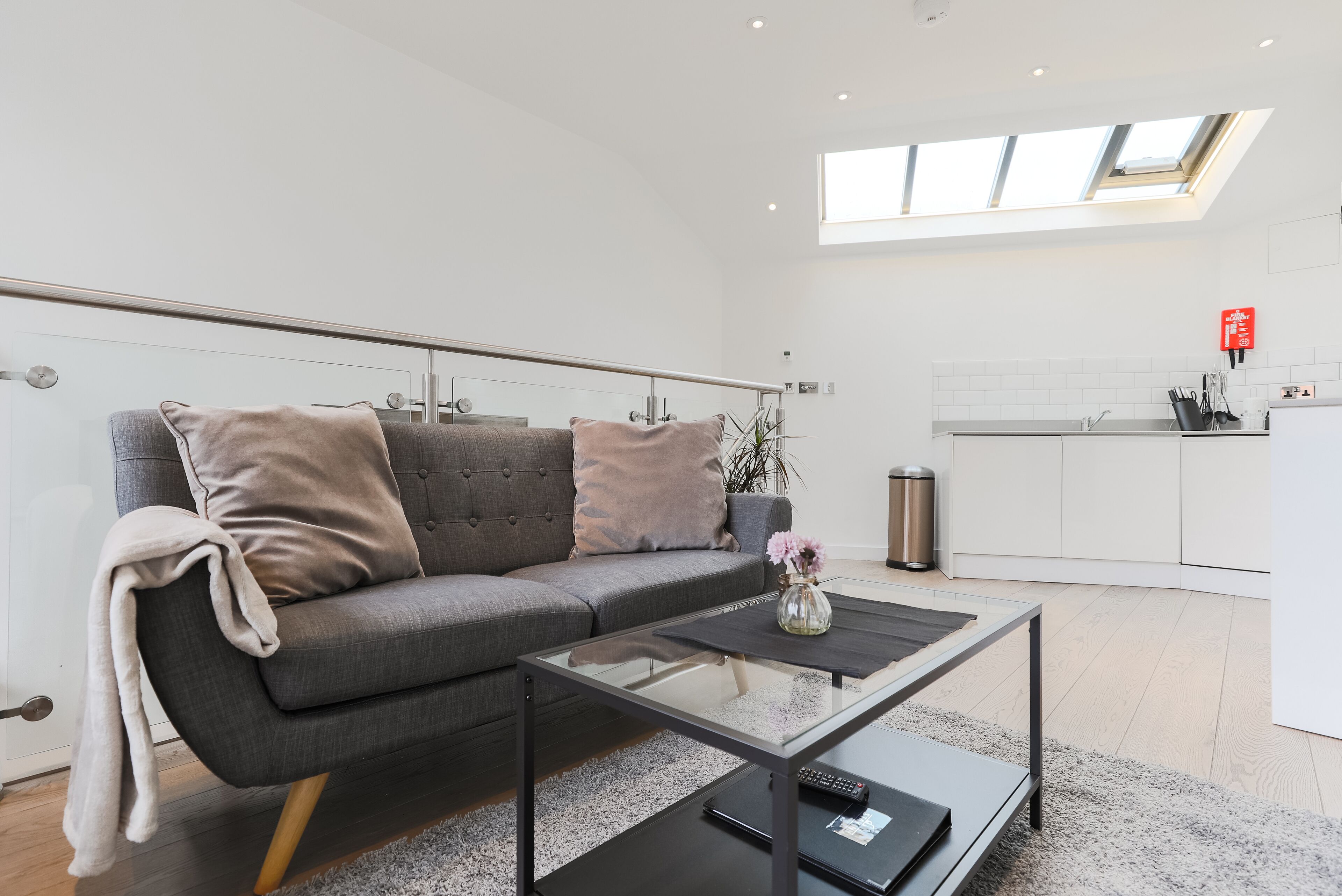 A modern living room with a grey sofa, a glass coffee table, and a kitchen in the background.