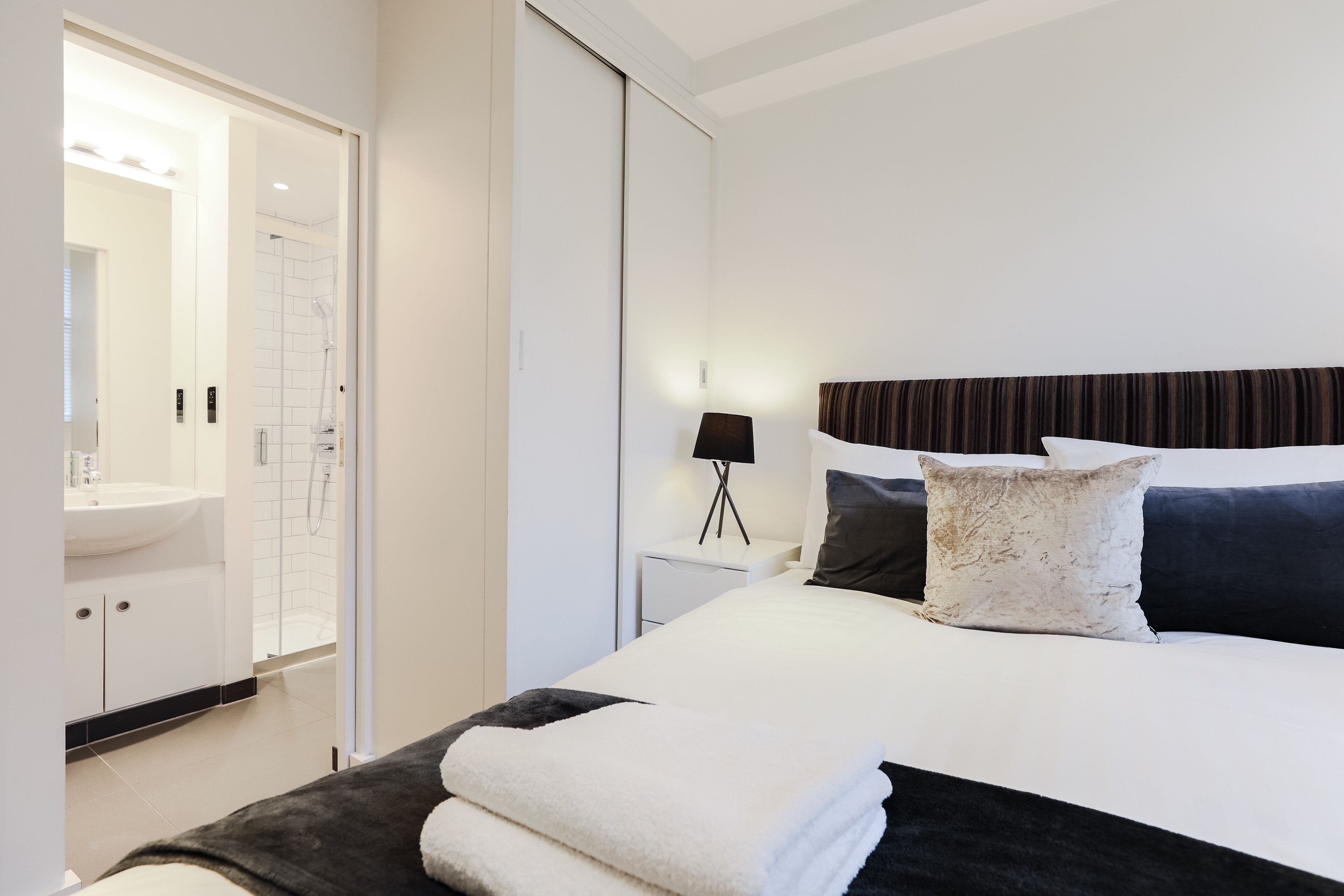 A modern hotel room with a large bed, a bedside table with a lamp, a bathroom with a sink and shower, and a wardrobe.