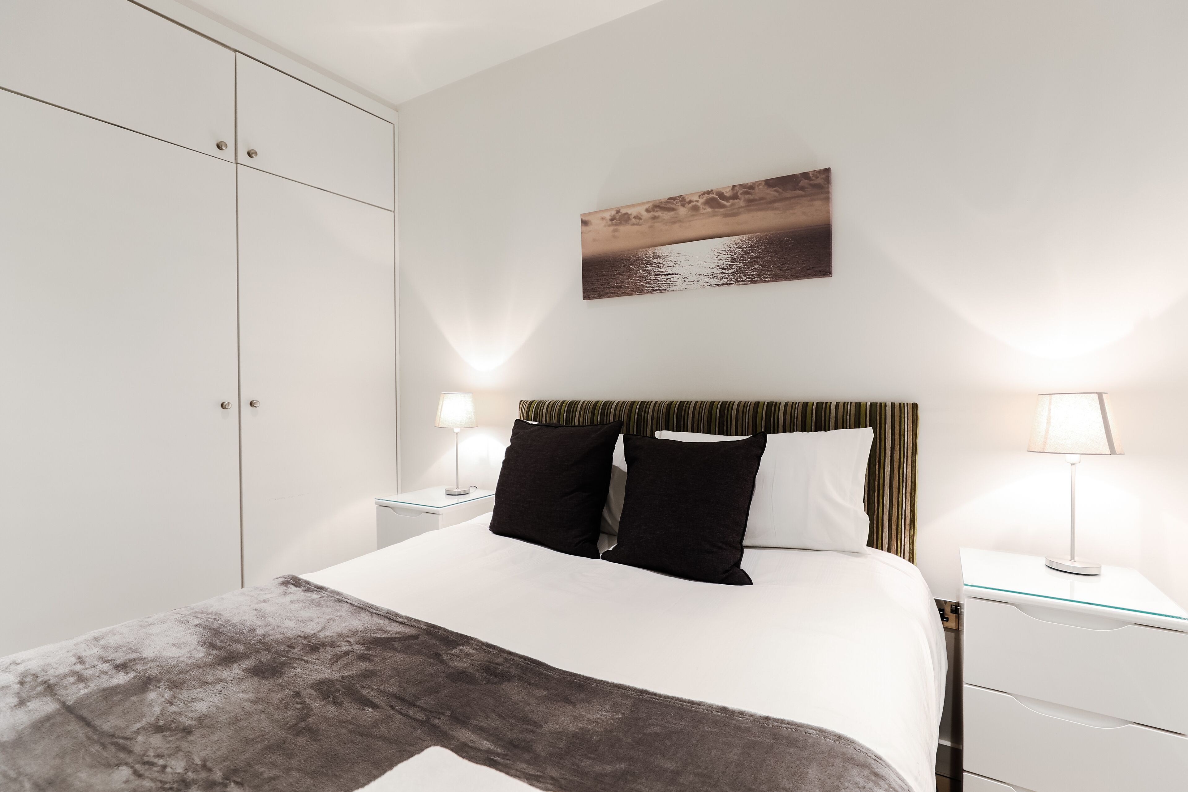 A modern bedroom with a large bed, two bedside tables with lamps, a wardrobe, and a landscape painting on the wall.