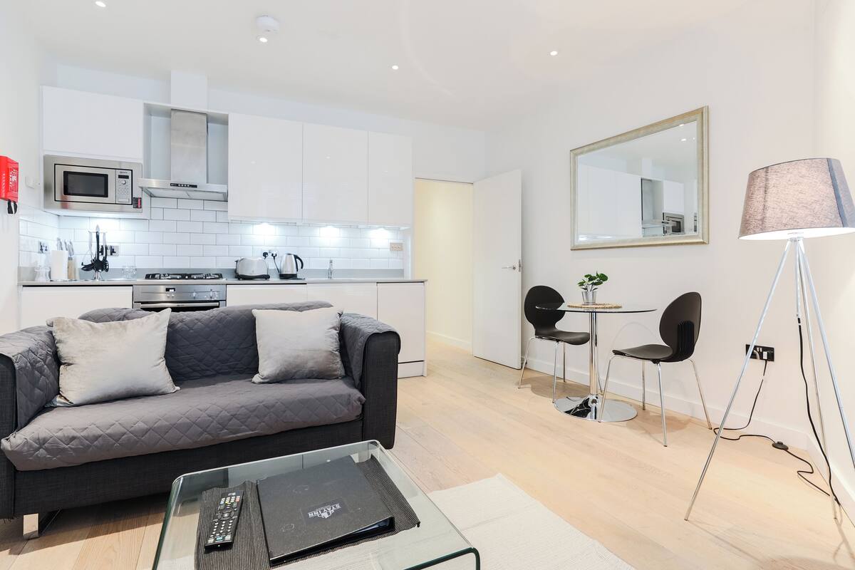 A modern living room with a grey sofa, a glass coffee table, and a dining area with two chairs.