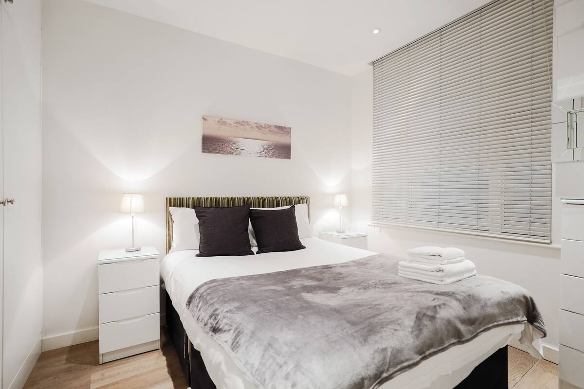 A modern bedroom with a bed, bedside lamps, a nightstand, a window with blinds, and a painting on the wall.