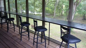 Un espacio exterior techado con una mesa larga y cuatro taburetes altos, con vistas a un área boscosa.