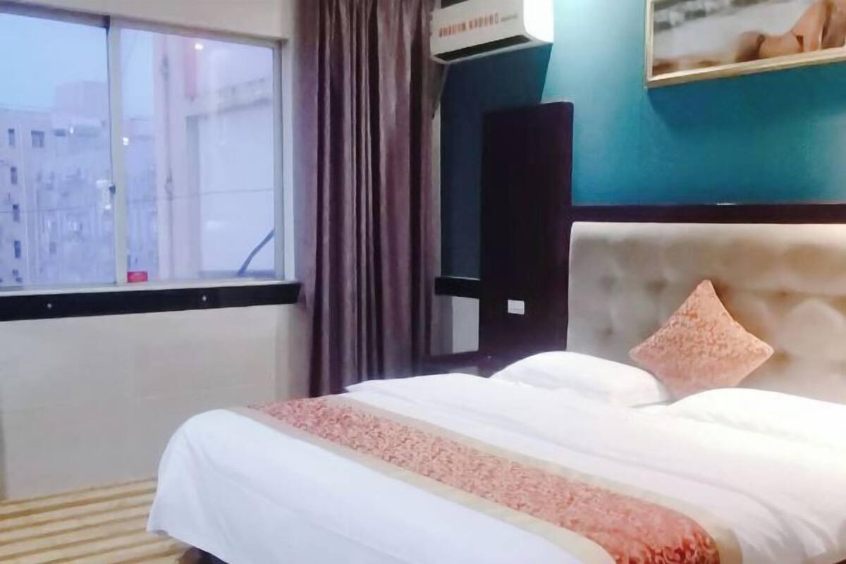 A hotel room with a bed, a window, curtains, an air conditioning unit, and a wall-mounted speaker.