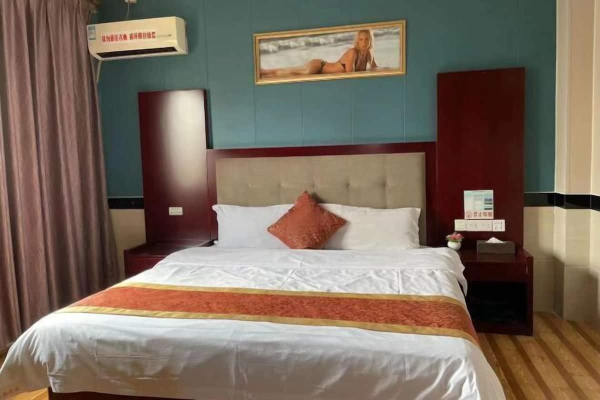 A hotel room with a large bed, a nightstand, a wall-mounted air conditioner, and a framed picture on the wall.