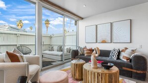 A modern living room with a grey sofa, a white armchair, and a round ottoman. There are three wooden side tables with decorative items. The room has large windows with a view of palm trees and a white fence. - Boho chic condo at the mid-century Ocotillo Lodge (Palm Springs)