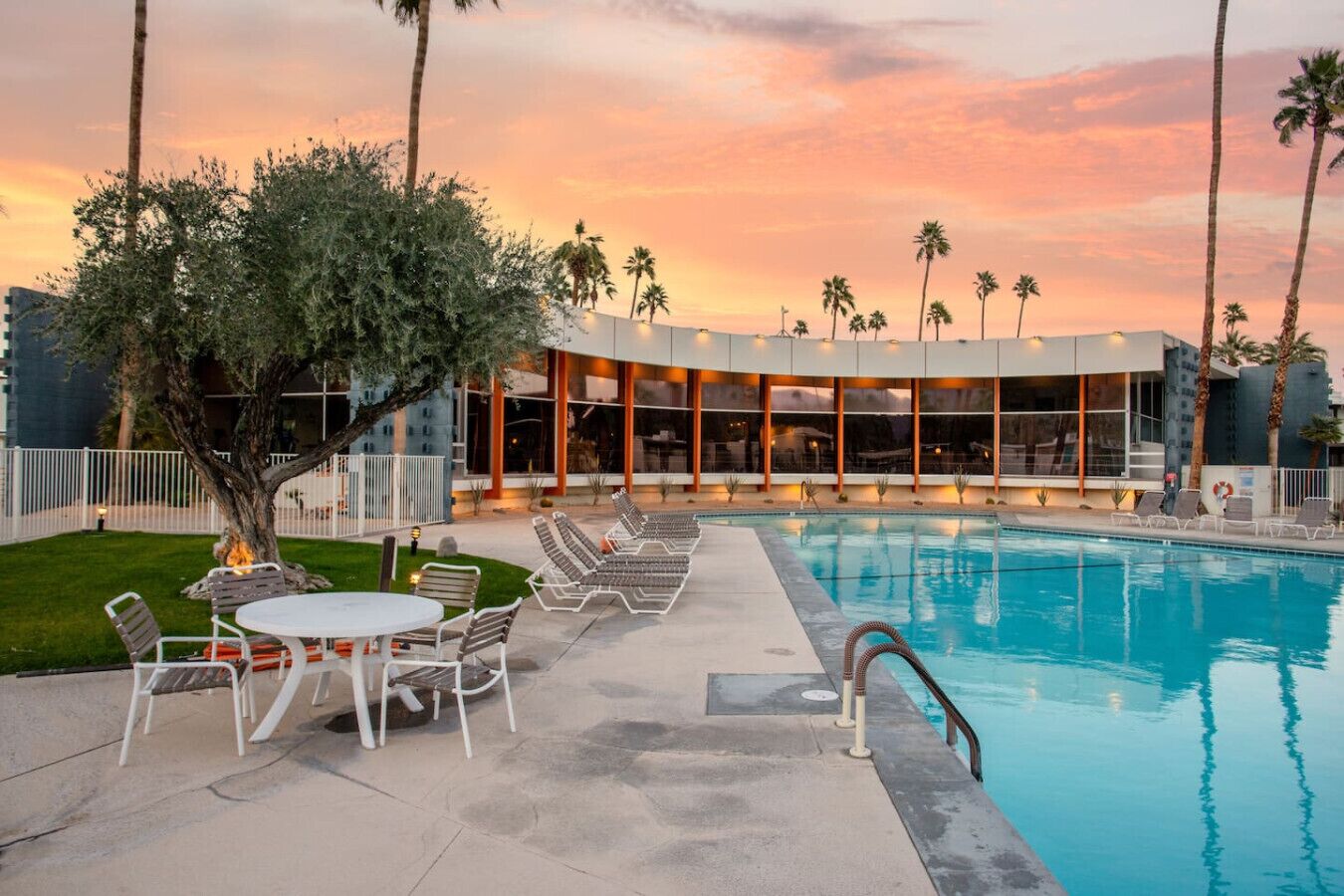 A pool area with lounge chairs and a table, surrounded by palm trees and a building with large windows.