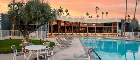 A pool area with lounge chairs and a table, surrounded by palm trees and a building with large windows.
