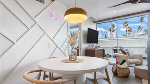A modern living room with a round dining table, wooden chairs, a ceiling fan, a TV, and a large window with a view of palm trees. - Boho chic condo at the mid-century Ocotillo Lodge (Palm Springs)