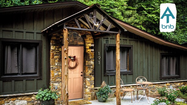 Una baita rustica con facciata in pietra, una porta in legno e un portico con sedie in metallo.