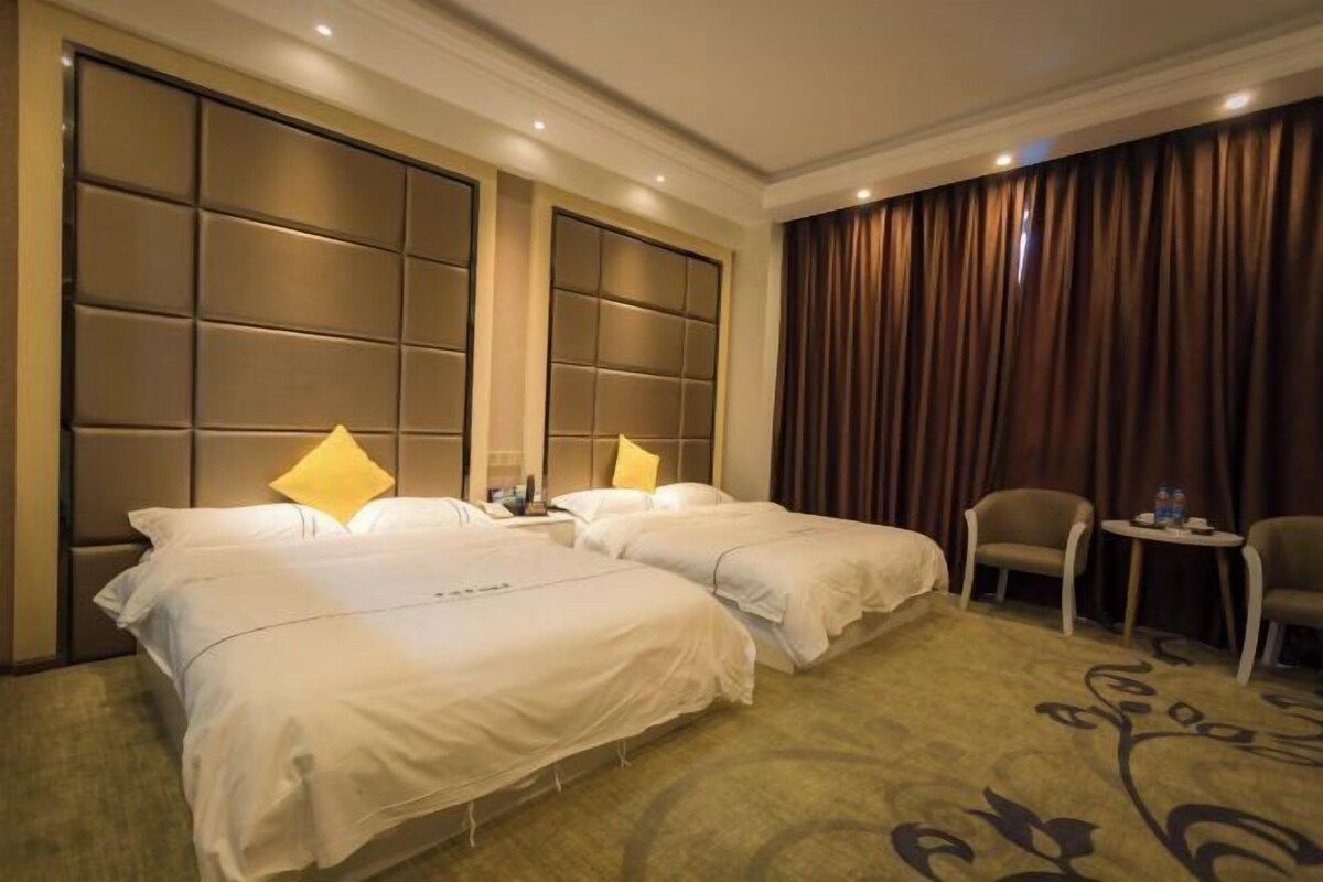 A hotel room with two beds, a large padded headboard, a small table with chairs, and a carpeted floor.
