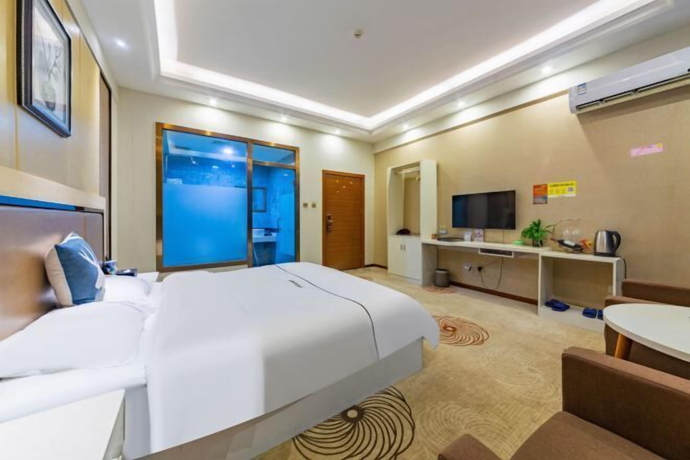 Quarto de hotel com cama, escrivaninha, televisão e banheiro visível por uma porta de vidro.