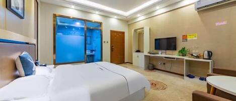 A hotel room with a bed, a desk, a TV, and a bathroom visible through a glass door.