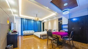 A modern hotel room with a wooden floor, a bed with a blue curtain, a dining area with a table and chairs, and a built-in wardrobe.