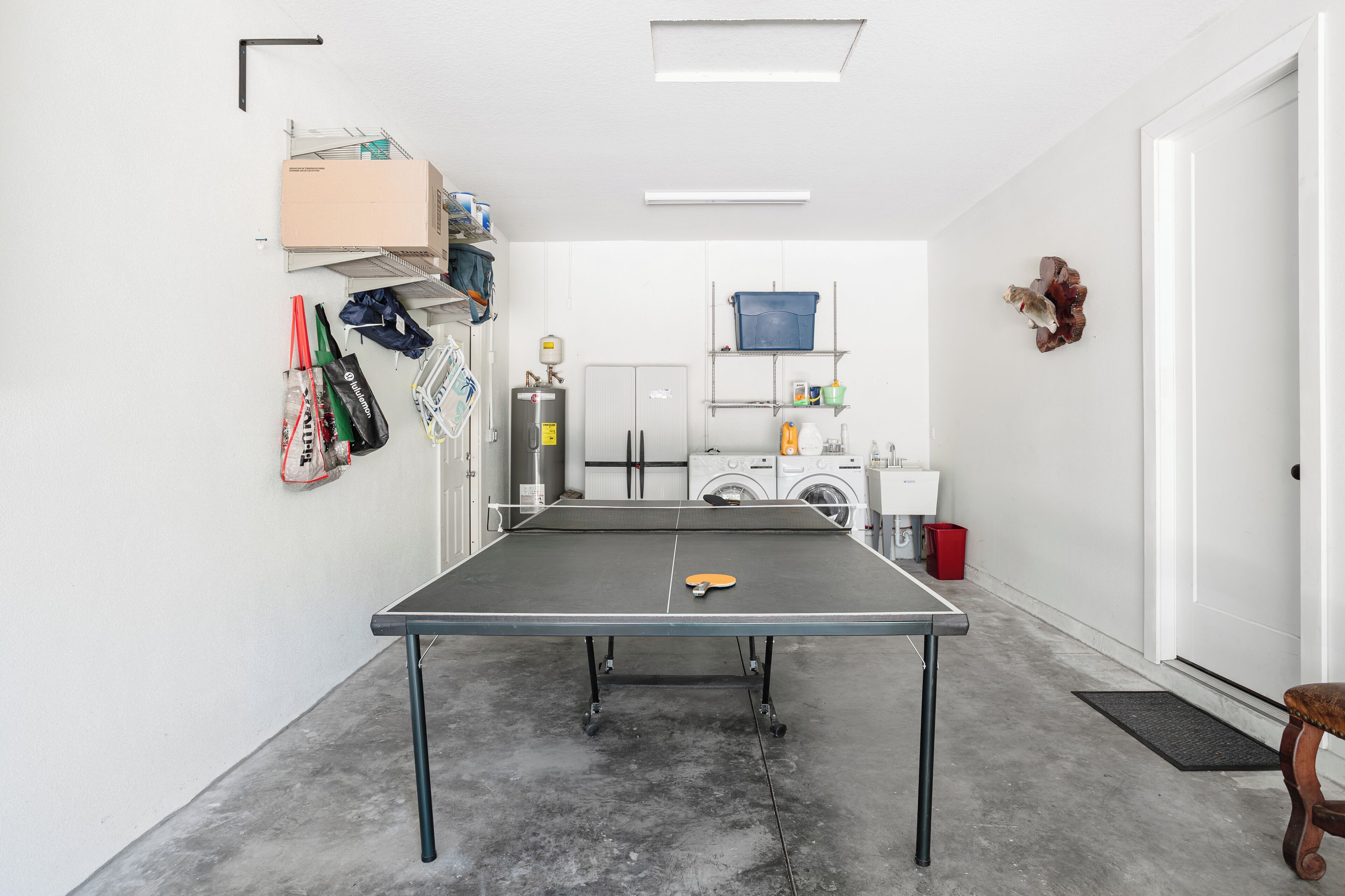 Une table de ping-pong dans une pièce où se trouvent un lave-linge, des étagères remplies de divers objets et une porte.