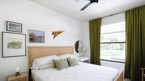 Ein Schlafzimmer mit Bett, Nachttisch, Spiegel, Fenster mit Vorhängen und Wandbildern.