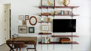 Ein Zimmer mit einem Holztisch, einem Sessel, einem Fernseher und Regalen mit Büchern und Dekoartikeln.