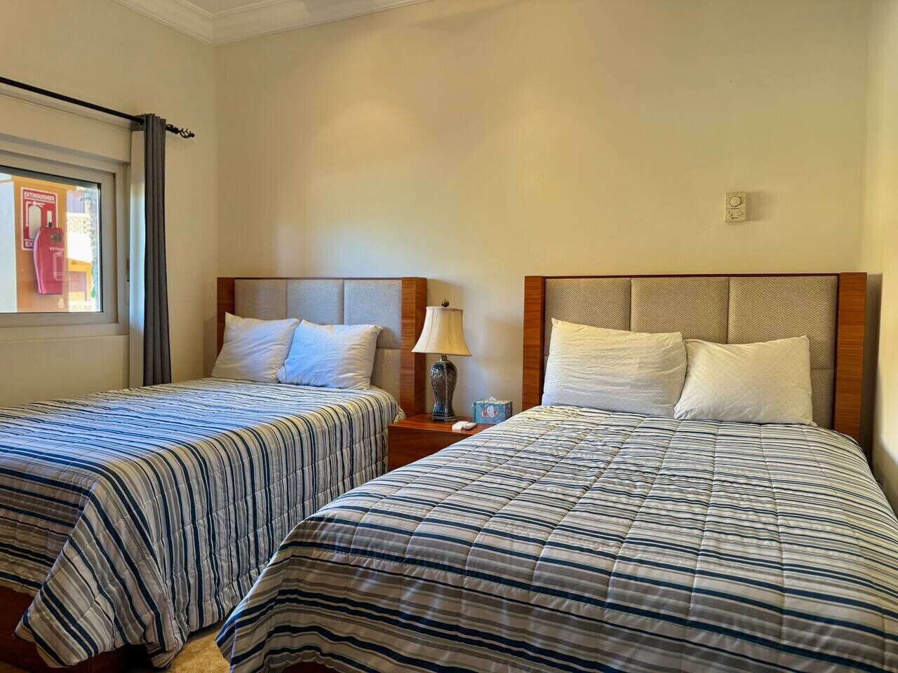 Two single beds with striped bedding, a nightstand with a lamp, and a window with a curtain.