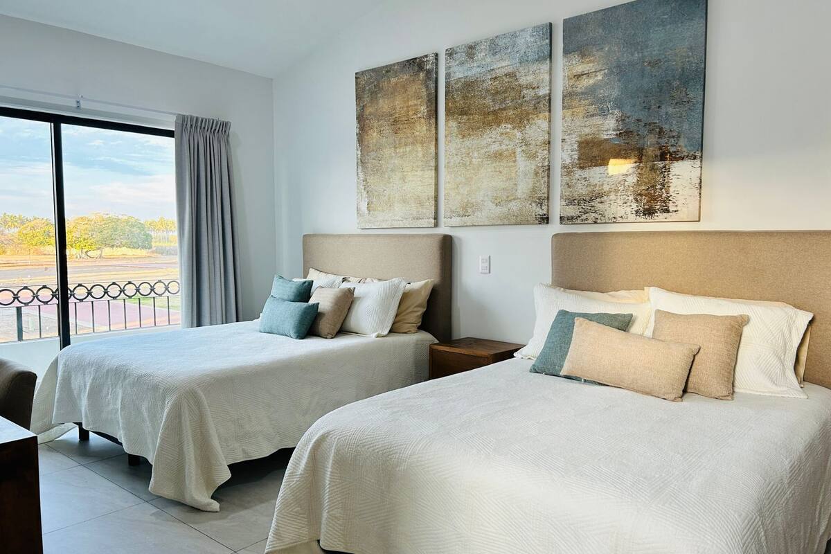 Una habitación de hotel con dos camas, un ventanal con vistas y tres cuadros abstractos en la pared.