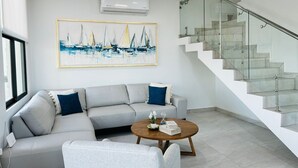 Una sala moderna con un sofá gris, una mesa de centro redonda y una escalera con barandillas de vidrio.