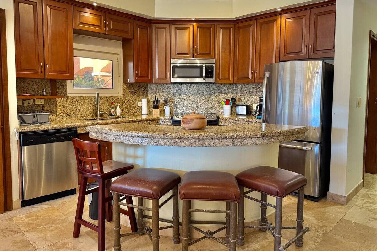 Una cocina con encimeras de granito, electrodomésticos de acero inoxidable, armarios de madera y una isla central con taburetes de bar.