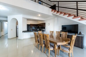 A spacious dining area with a large wooden table and chairs, a kitchen in the background, and a staircase leading to an upper level.