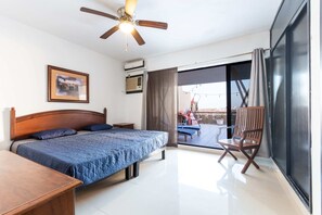 A bedroom with a bed, a wooden nightstand, a ceiling fan, an air conditioning unit, and a large window with a view of outdoor seating.