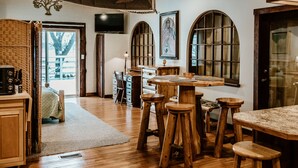 A spacious room with wooden flooring, a large wooden table, and several wooden stools. There is a television mounted on the wall, a door leading outside, and a window with a view of the outside.