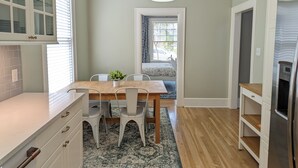 Een keuken met een houten tafel, witte stoelen, een hangende lamp en uitzicht door een raam.