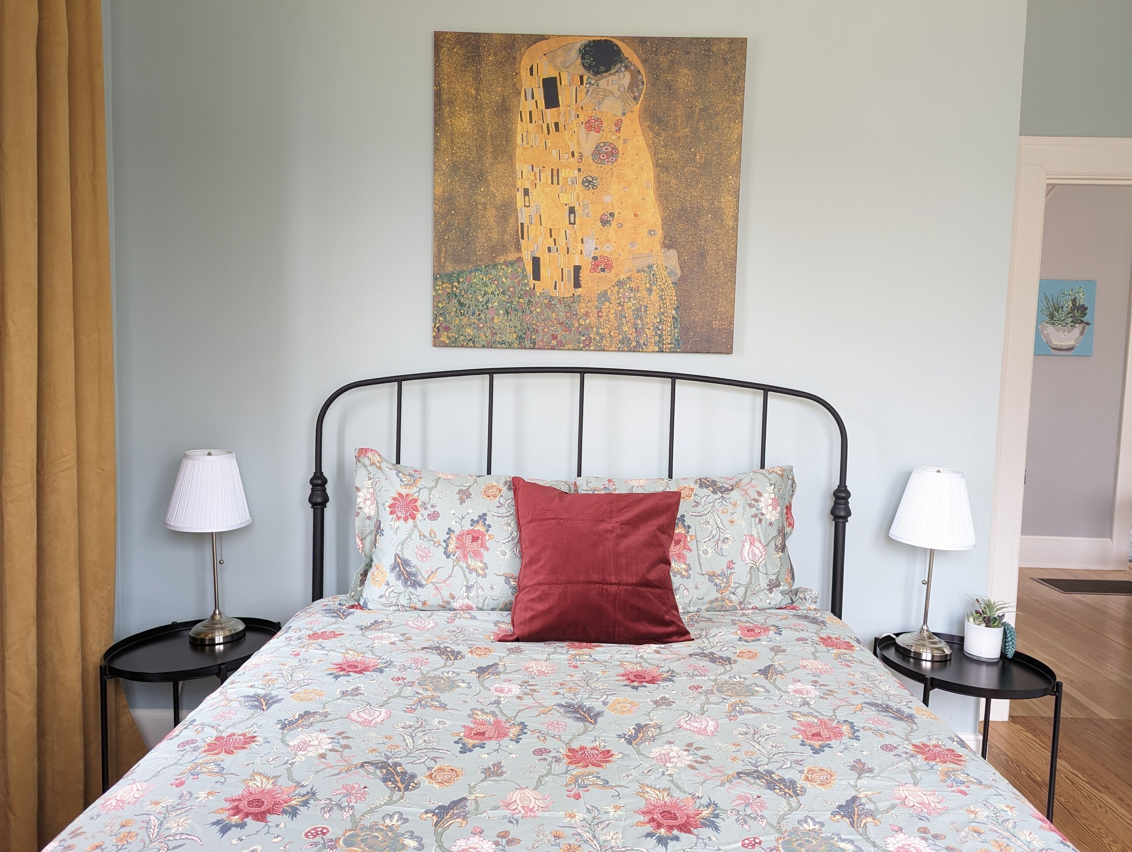Ein Schlafzimmer mit einem Bett, floralem Bettzeug, zwei Nachttischen mit Lampen, einem roten Kopfkissen und einem Gemälde über dem Bett.