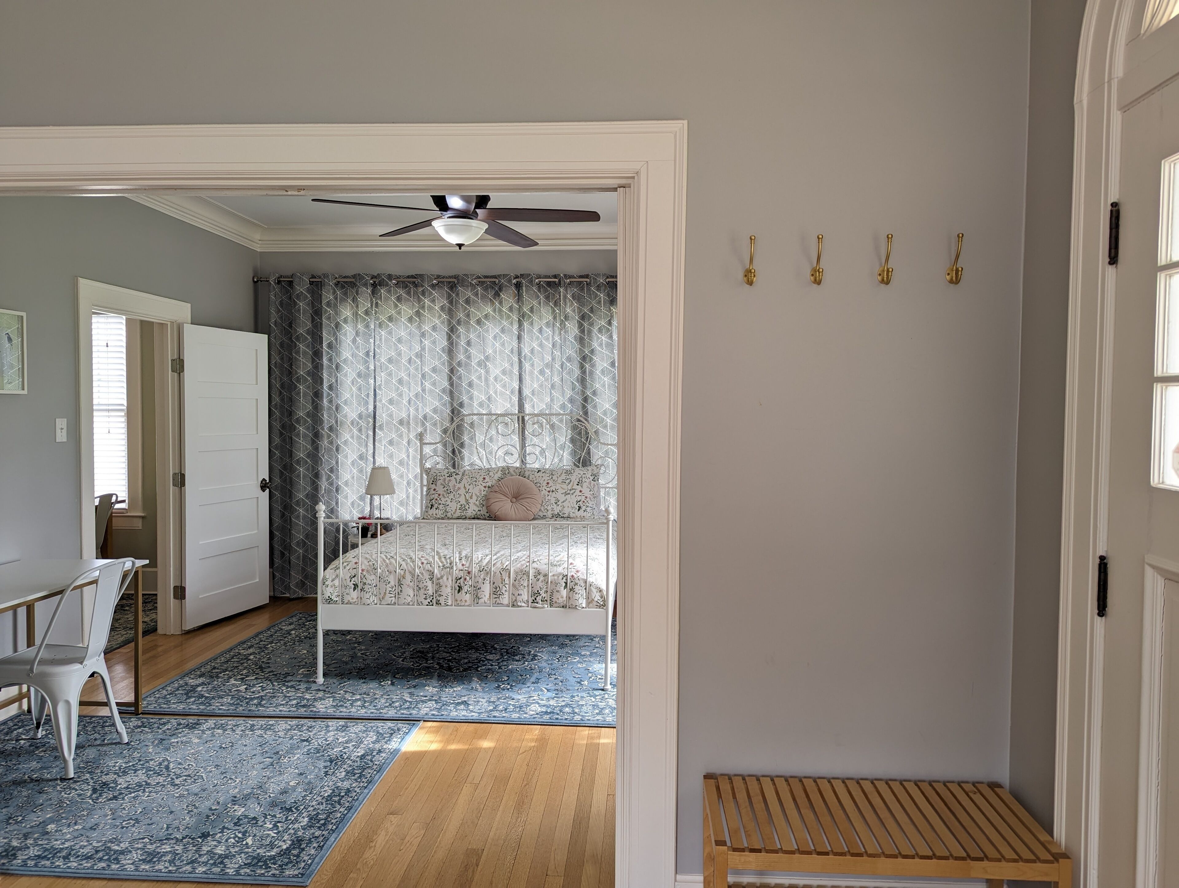 A bedroom with a bed, a desk, a chair, a wooden bench, a ceiling fan, and patterned curtains.