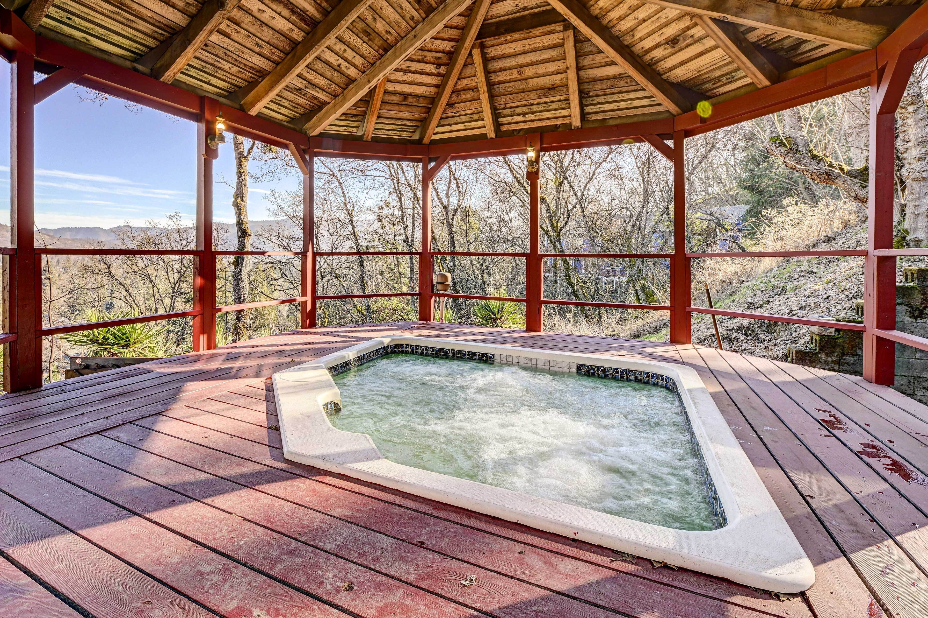 A hot tub on a wooden deck with a wooden pergola overhead, surrounded by trees and a mountainous backdrop.